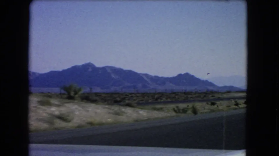 1966 Nevada home-movie frame from a moving car, showing open desert, scattered shrubs, road foreground, and distant mountains.