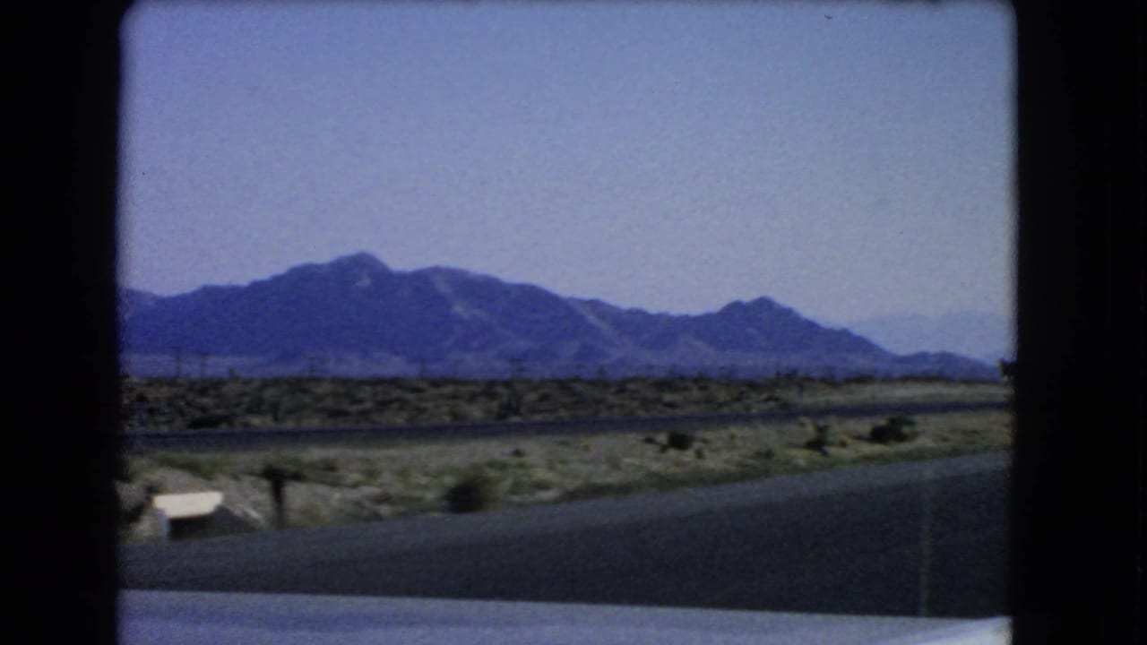 Las Vegas-area 1966 8mm road footage showing desert scrub, paved road, mountain backdrop, and a small light roadside object.