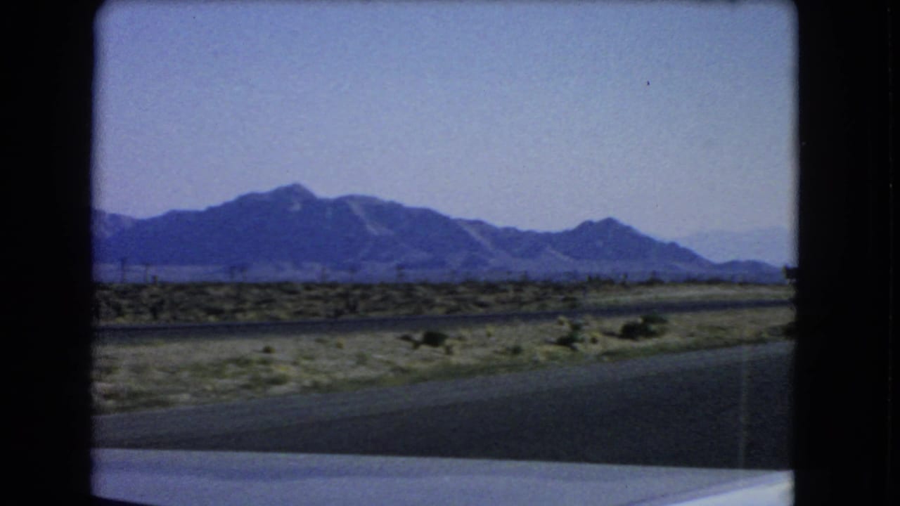 1966 Las Vegas-area 8mm frame from a moving vehicle showing desert roadside, scrub, utility poles, and distant mountains.