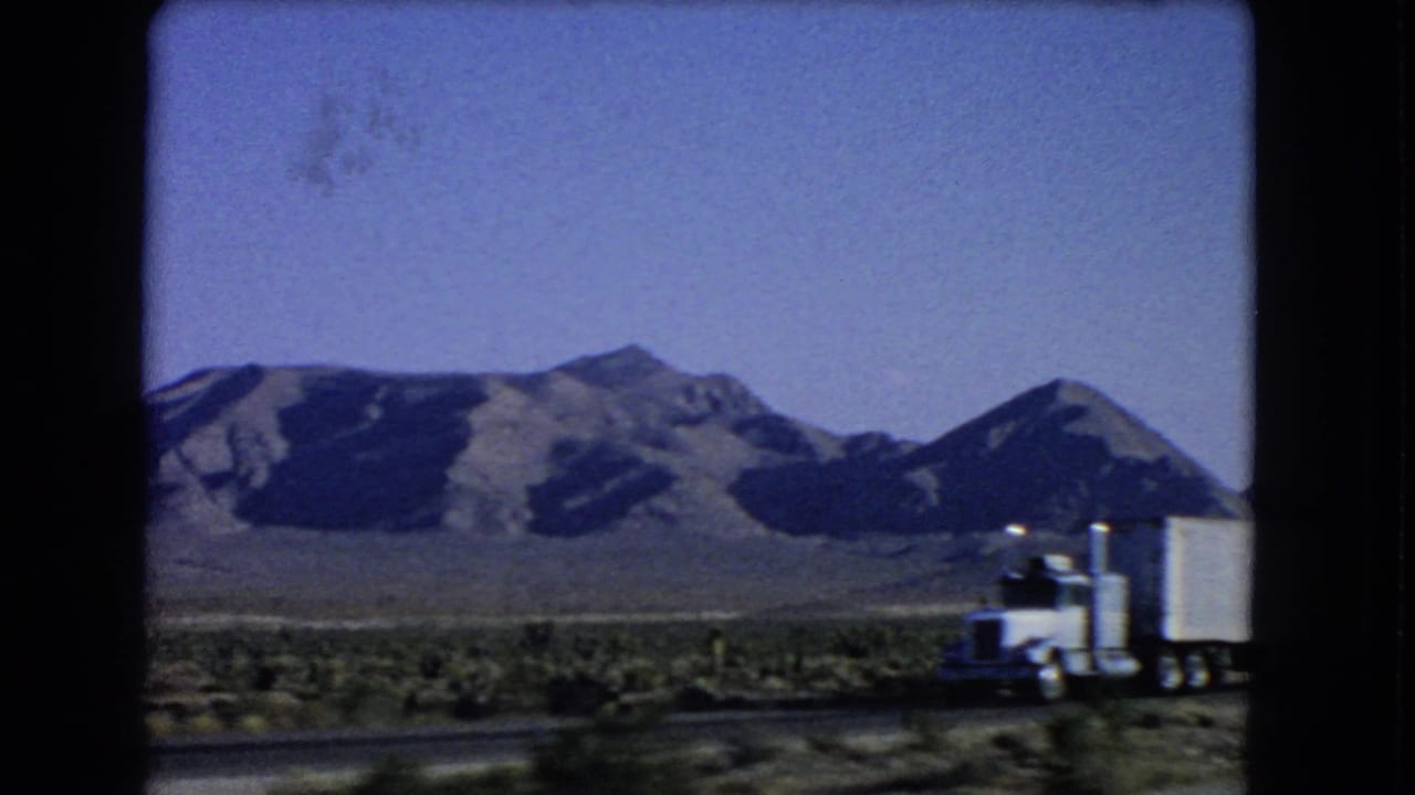 A white semi-truck moves along a Las Vegas-area desert highway with rugged mountains and sparse vegetation behind it.