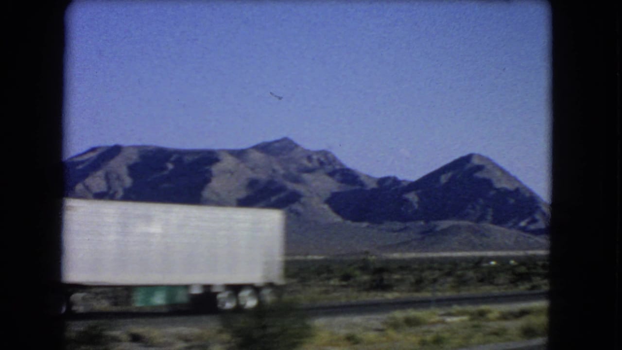 The side of a white trailer crosses the foreground as the moving camera passes desert mountains near Las Vegas in 1966.