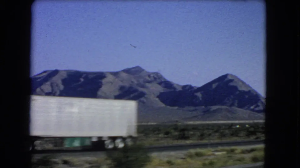 The side of a white trailer crosses the foreground as the moving camera passes desert mountains near Las Vegas in 1966.