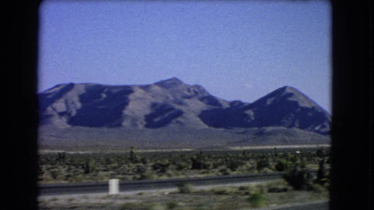Desert mountains and roadside brush seen from a moving highway vehicle.