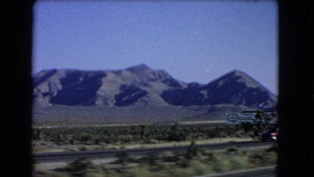 1966 Nevada highway view showing mountains, desert vegetation, road surface, and a partially visible roadside commercial sign.