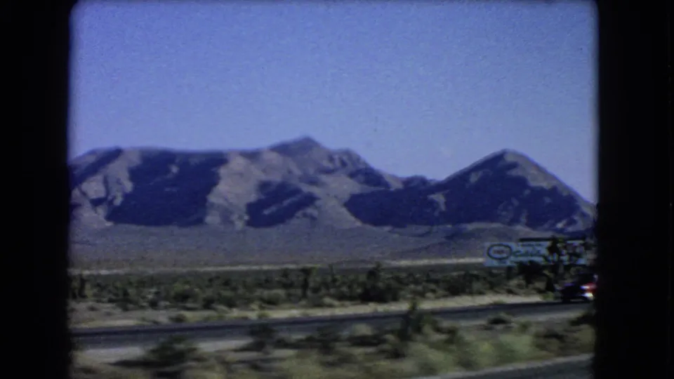 1966 Nevada highway view showing mountains, desert vegetation, road surface, and a partially visible roadside commercial sign.