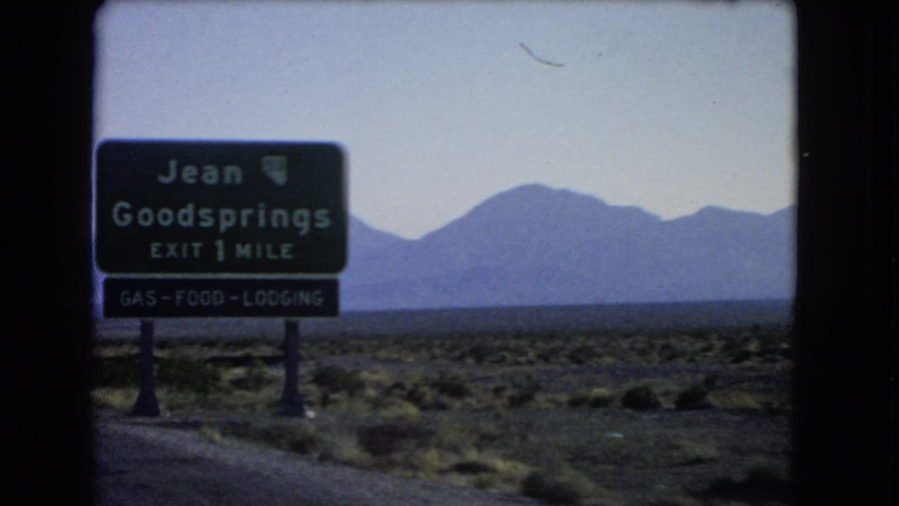 Highway sign for Jean and Goodsprings beside desert road and distant mountains.