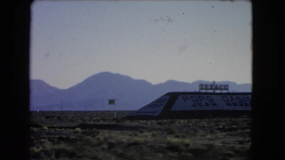 1966 Nevada desert roadside view with Texaco and Pops Oasis signage, low building roofline, scrubland, and mountains.