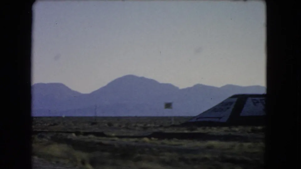 1966 Nevada road-trip frame showing part of a roadside structure, desert vegetation, small signs, and a mountain range under a pale sky.