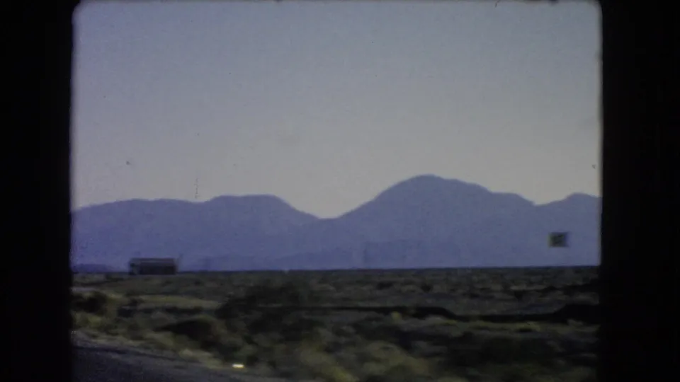 1966 Nevada desert view with scrubland, pale sky, distant mountains, and a small roadside structure near the horizon.