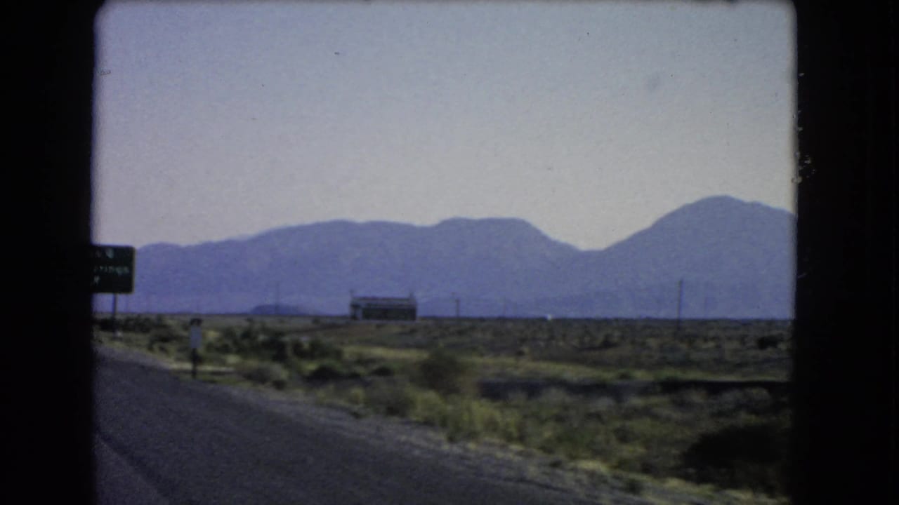 Passenger-window view of a Nevada desert road with roadside signs, a low building, utility poles, and distant mountains.