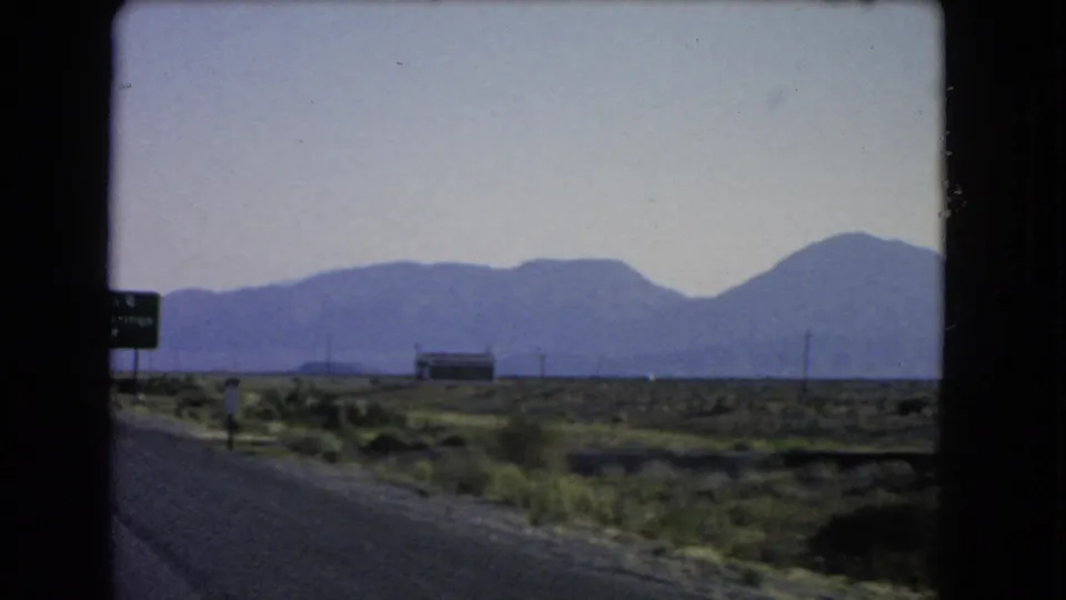 Passenger-window view of a Nevada desert road with roadside signs, a low building, utility poles, and distant mountains.