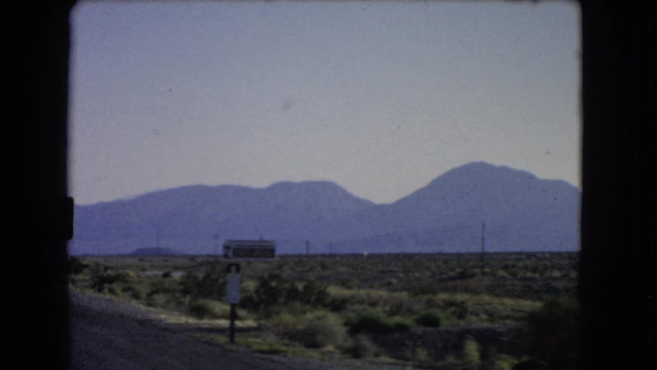 1966 Nevada road-trip frame showing a roadside signpost, sparse desert vegetation, utility poles, and a mountain backdrop.