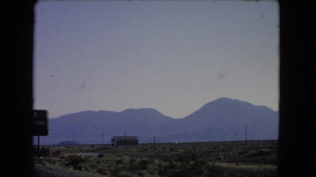 Soft 8mm view across Nevada desert scrub toward a low roadside structure, utility poles, and blue mountains on the horizon.
