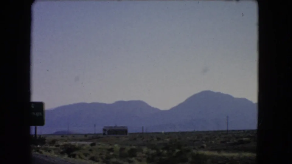 Soft 8mm view across Nevada desert scrub toward a low roadside structure, utility poles, and blue mountains on the horizon.