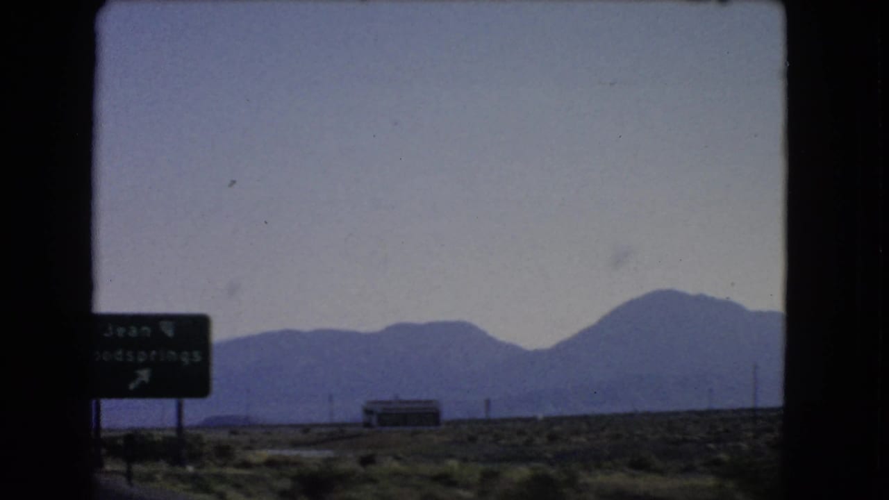 1966 Nevada roadside view from a moving car, with a Jean and Goodsprings sign entering frame against desert and mountains.