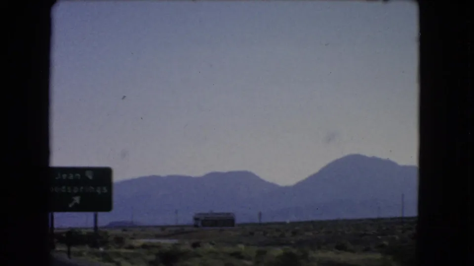 1966 Nevada roadside view from a moving car, with a Jean and Goodsprings sign entering frame against desert and mountains.