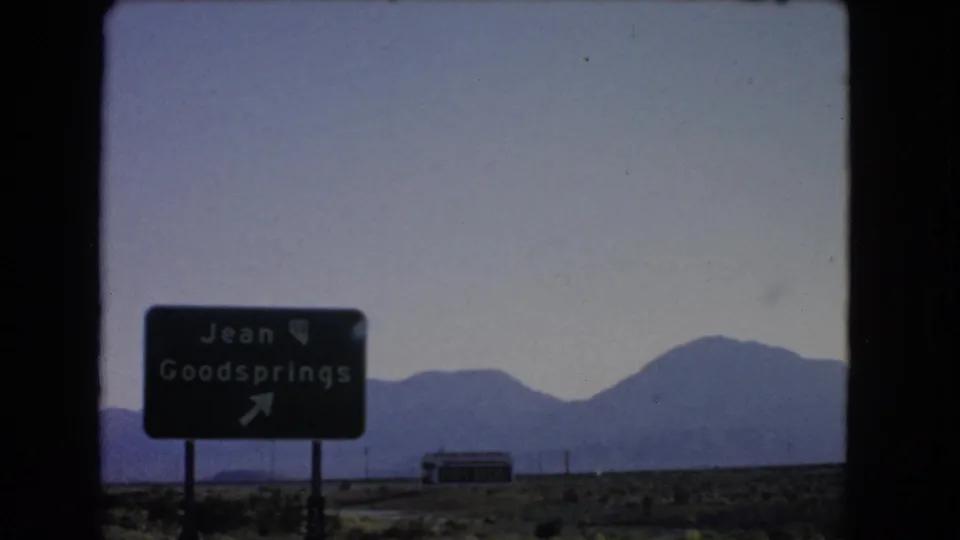 Jean and Goodsprings sign in 1966 Nevada, seen through a car window with desert scrub, mountains, and a small building beyond.