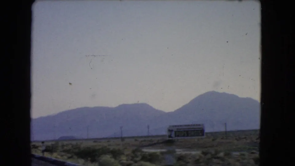 1966 Nevada desert roadside with a Pops Oasis billboard, utility poles, scrubland, and hazy mountains beyond the car window.