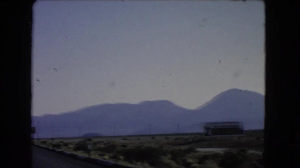 1966 Las Vegas-area 8mm frame showing desert roadside, a low distant structure, pale sky, and blue mountains beyond the highway edge.