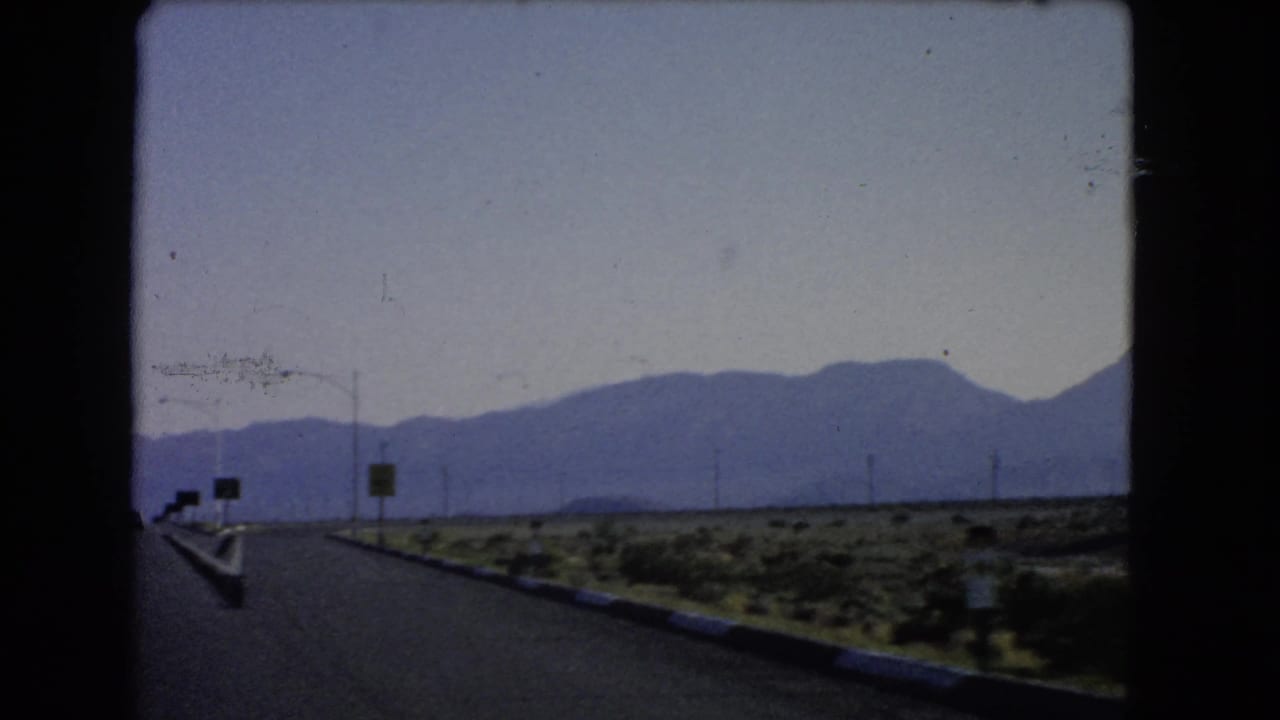 Open highway leading toward distant blue mountains with roadside signs and desert scrub.