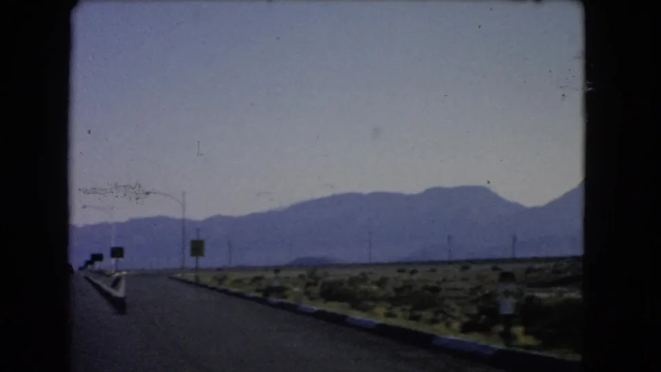 1966 Nevada road-travel frame with a wide highway, roadside signs, sparse desert scrub, utility poles, and hazy mountains ahead.