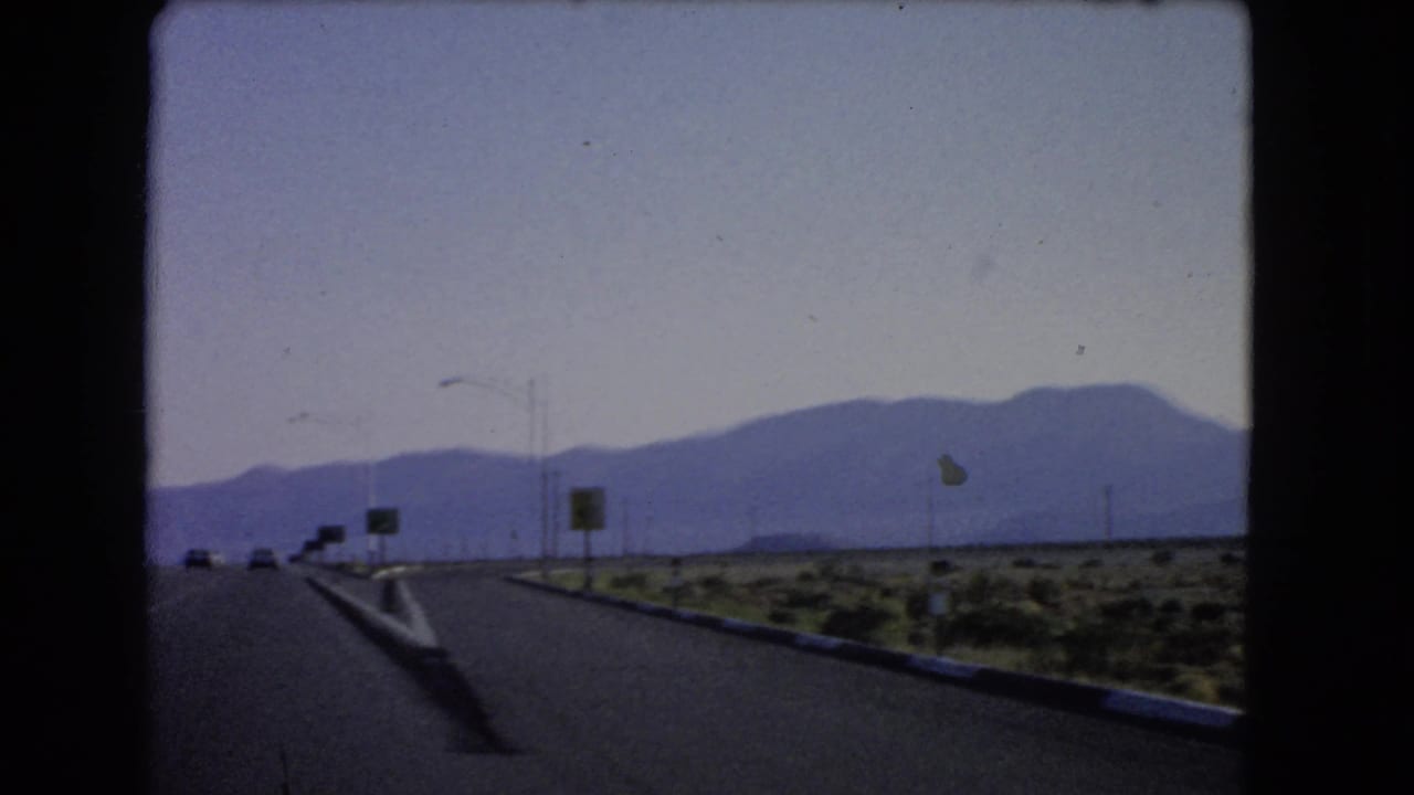 1966 Las Vegas-area highway view from a moving vehicle, showing distant cars, road signs, utility poles, desert brush, and mountains.