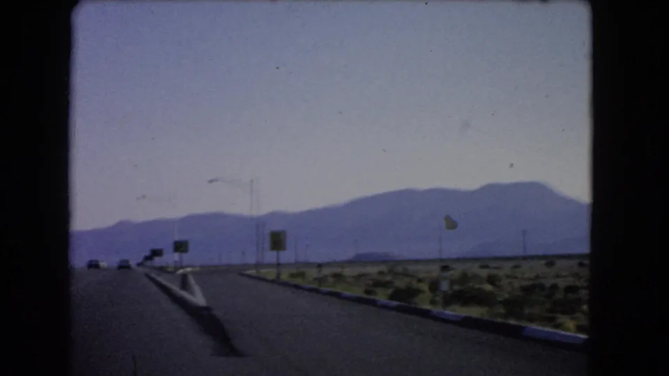 1966 Las Vegas-area highway view from a moving vehicle, showing distant cars, road signs, utility poles, desert brush, and mountains.