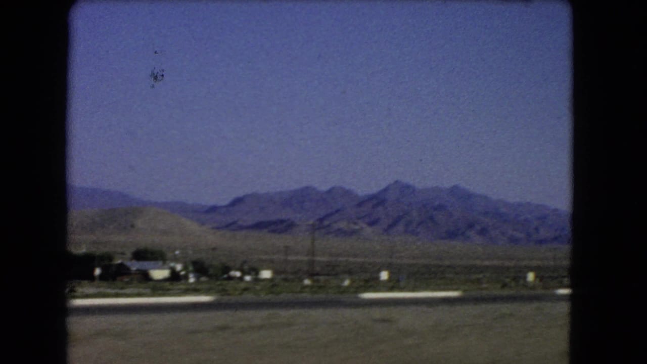 1966 Nevada desert frame showing low roadside buildings or settlement, open ground, utility poles, and a rugged mountain range.