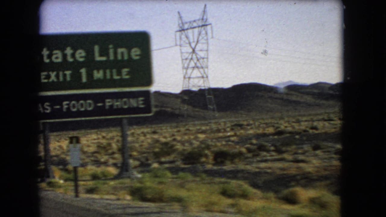 Green state-line exit sign beside desert hills and power lines.