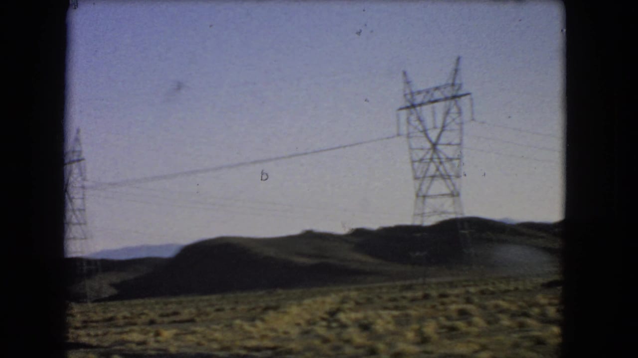 Power pylons and overhead lines stretch across low desert hills in vintage 8mm Nevada travel footage.