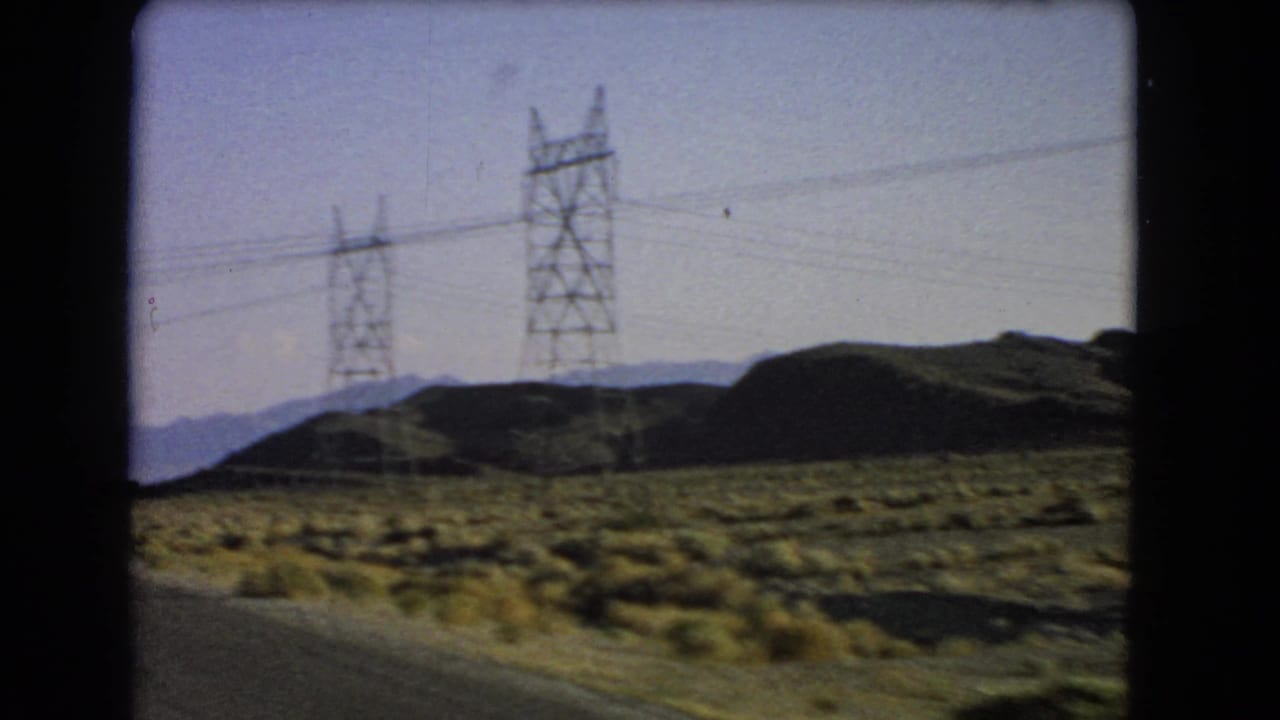 Roadside desert view with utility towers, power lines, scrubland, hills, and distant mountains.
