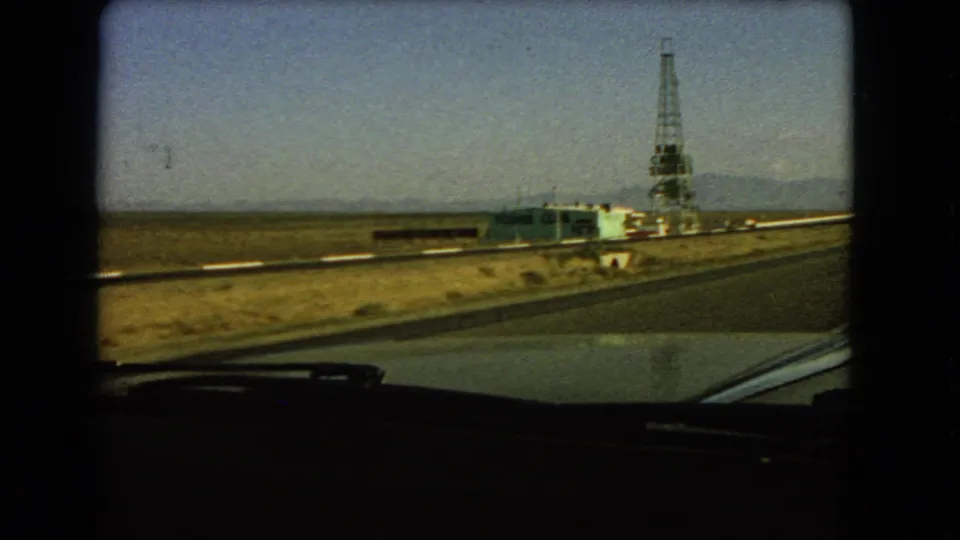 Las Vegas-area roadside view from inside a moving car, showing a low structure and tall derrick-like tower in the desert.