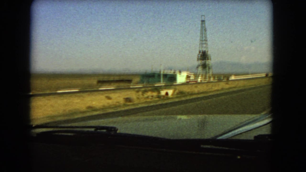 1966 Las Vegas-area 8mm view from a moving car toward a roadside tower, low building, dry valley, and distant mountains.