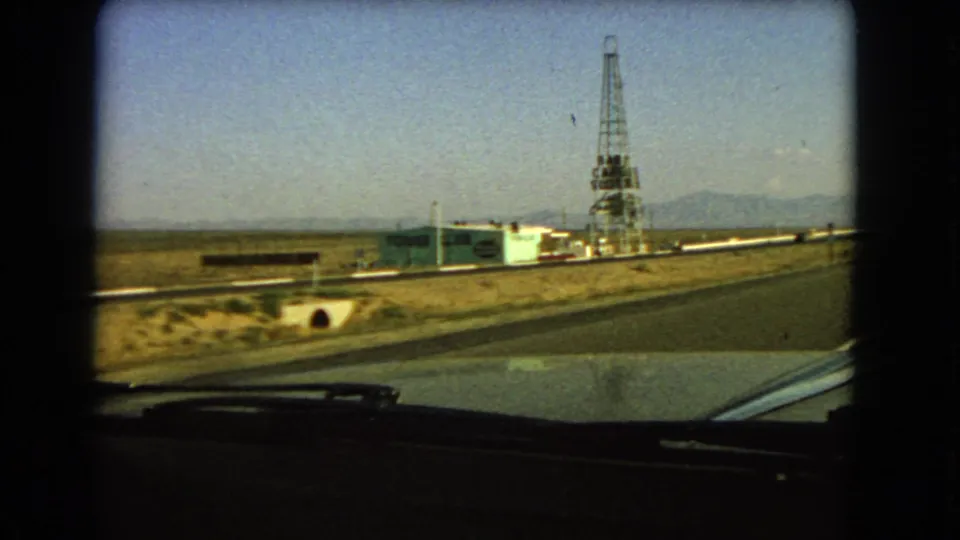 Car-window view in 1966 Las Vegas, Nevada, showing a tower, roadside facility, open highway edge, dry terrain, and mountain horizon.