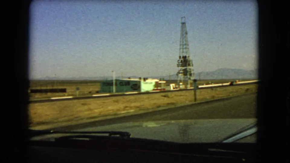 1966 8mm Las Vegas-area road view with a green roadside building, tall tower, open valley floor, and mountains beyond.
