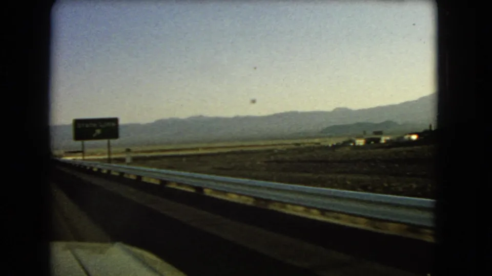 1966 desert highway view with a State Line sign, metal guardrail, open land, and mountain range in the distance.