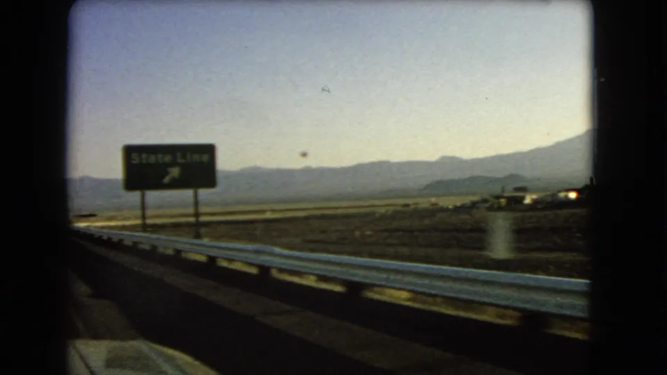 Passenger view of a State Line highway sign in the Nevada desert, with a guardrail and distant mountains under a pale sky.