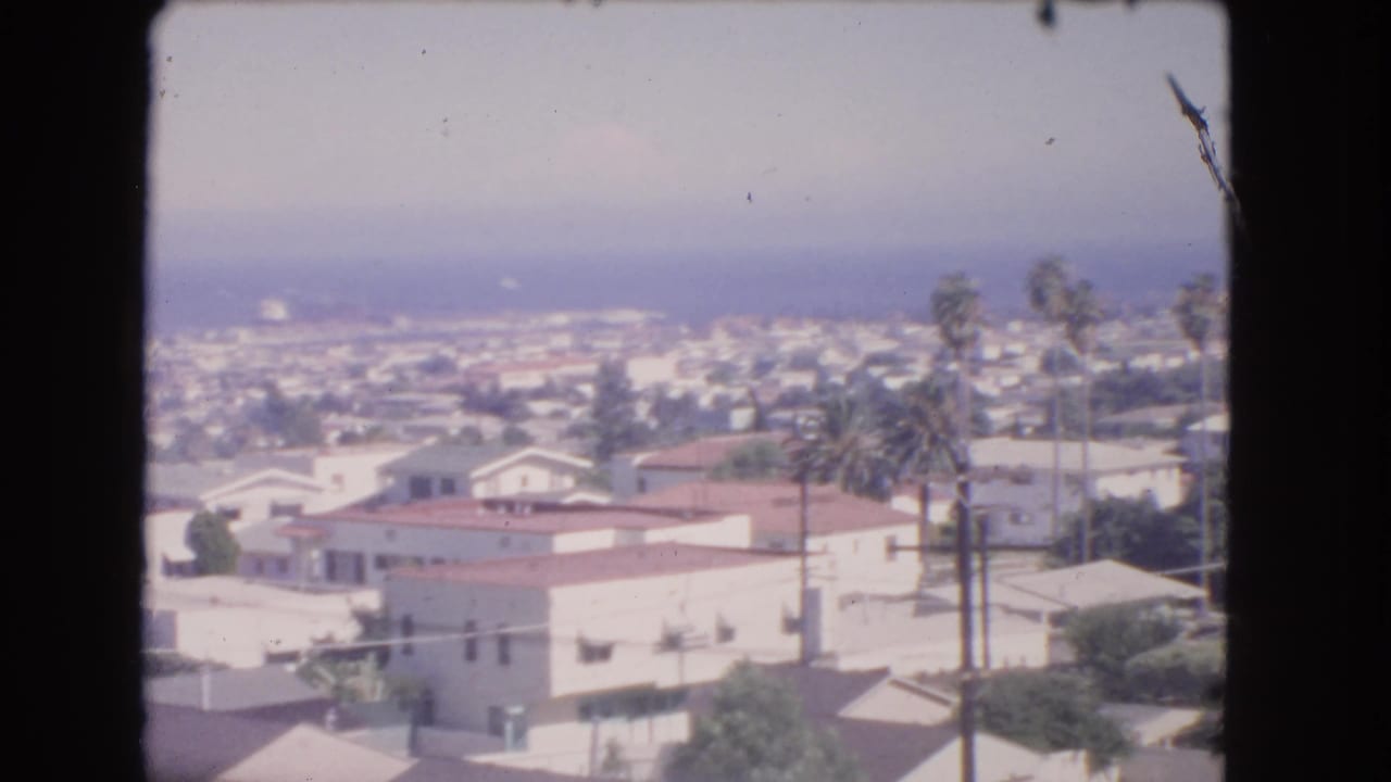Distant Los Angeles residential view with stucco houses, red roofs, palm trees, and ocean haze.