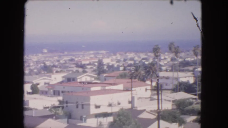 1966 Los Angeles home-movie frame showing white houses, red-tiled roofs, tall palms, and a hazy coastal horizon.