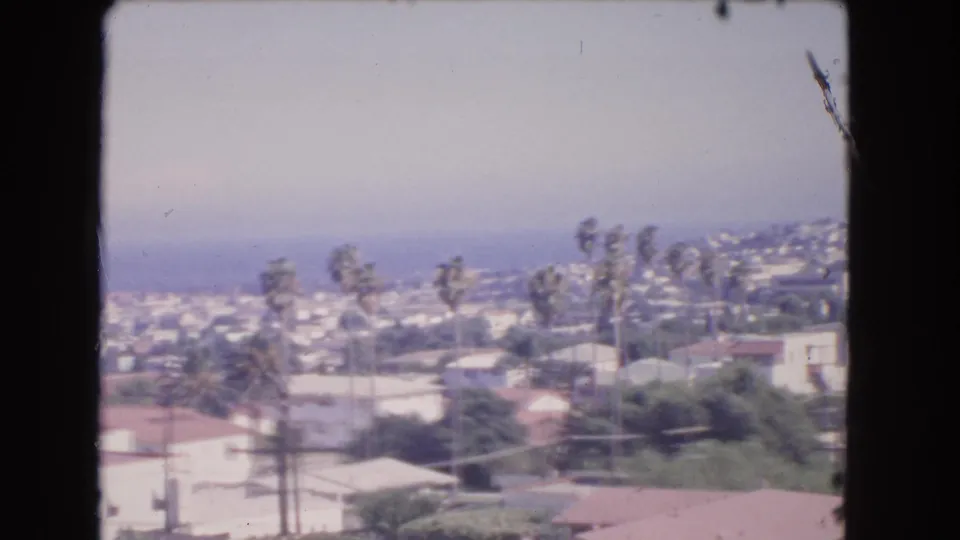 1966 Los Angeles frame with hillside residences, palms, utility poles, and a soft blue-gray coastal distance.