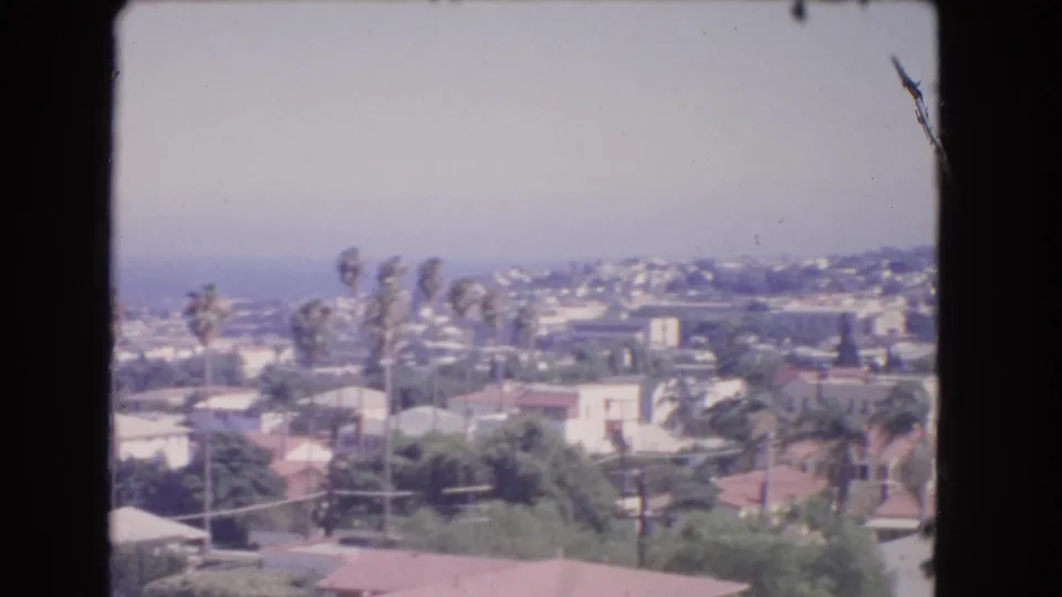 1966 Los Angeles home-movie frame showing rooftops, palms, hillside housing, and dark borders from the shooting position.