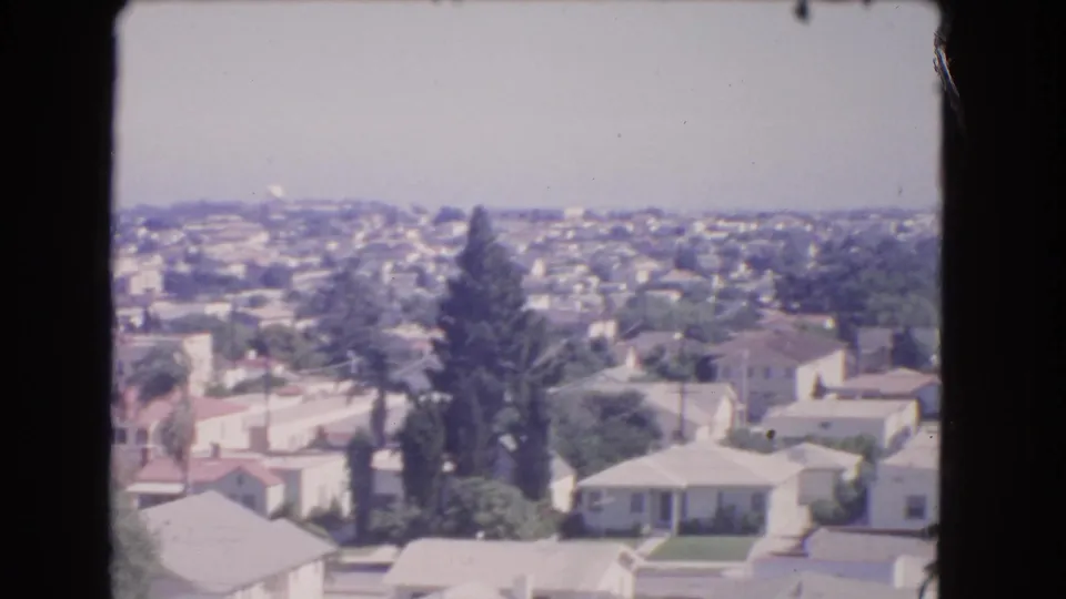 1966 Los Angeles elevated view showing residential blocks, palms, pale rooftops, and a hazy cityscape beyond.