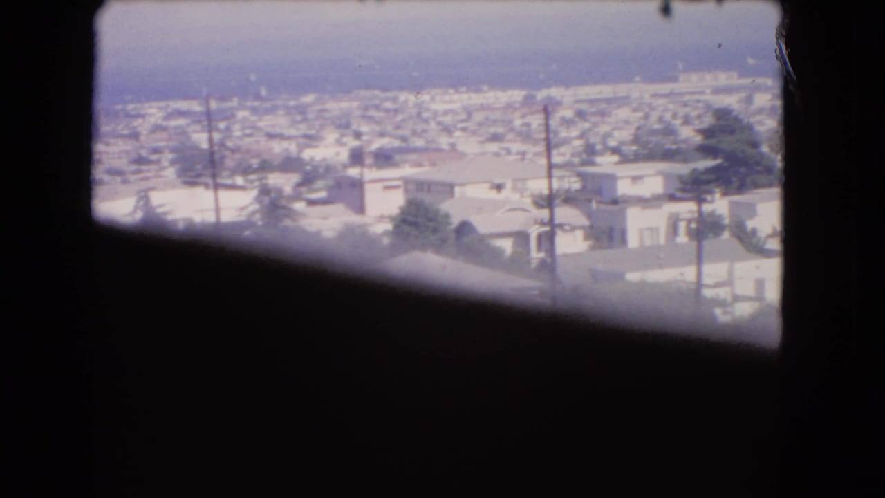 Dark window frame overlooking rooftops, utility poles, trees, and distant water.