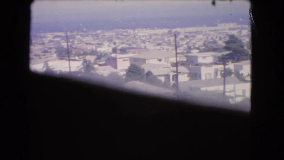 1966 Los Angeles home-movie frame looking through a dark window opening toward pale rooftops, utility poles, trees, haze, and distant water.