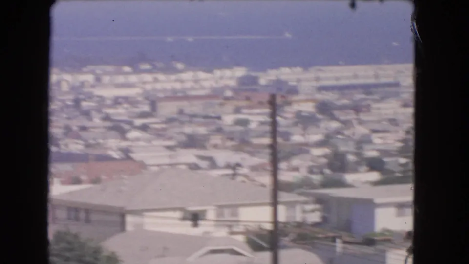 Soft 8mm frame from Los Angeles showing rooftops, a foreground utility pole, hillside structures, trees, and distant blue water.