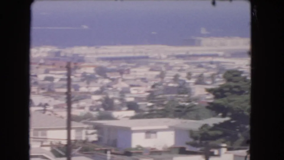 1966 Los Angeles home-movie still with pale rooftops, a vertical utility pole, trees, low-rise buildings, haze, and a distant coastal backdrop.