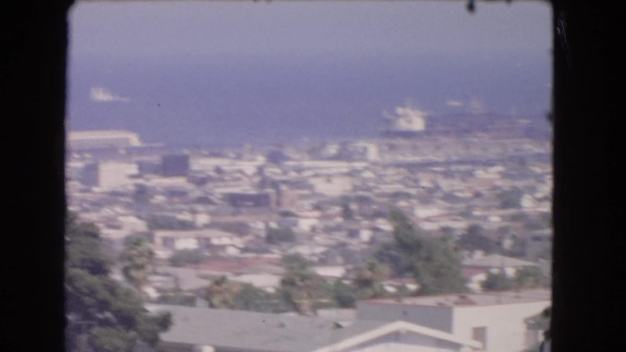 High-angle 1966 Los Angeles coastal city view showing dense rooftops, palm trees, blue ocean, and distant ships offshore.