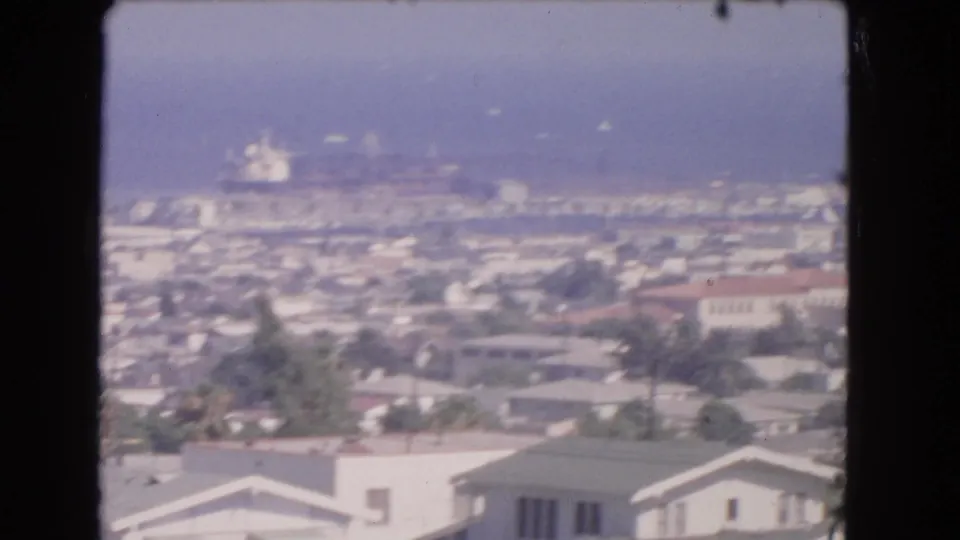 1966 home-movie frame of a Los Angeles coastal cityscape, with homes, palms, harbor structures, ocean, and a ship in the haze.