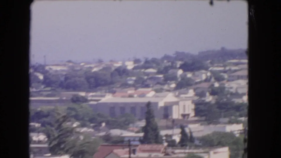 1966 Los Angeles frame showing a hillside residential area, palms, rooftops, and a pale building under hazy light.