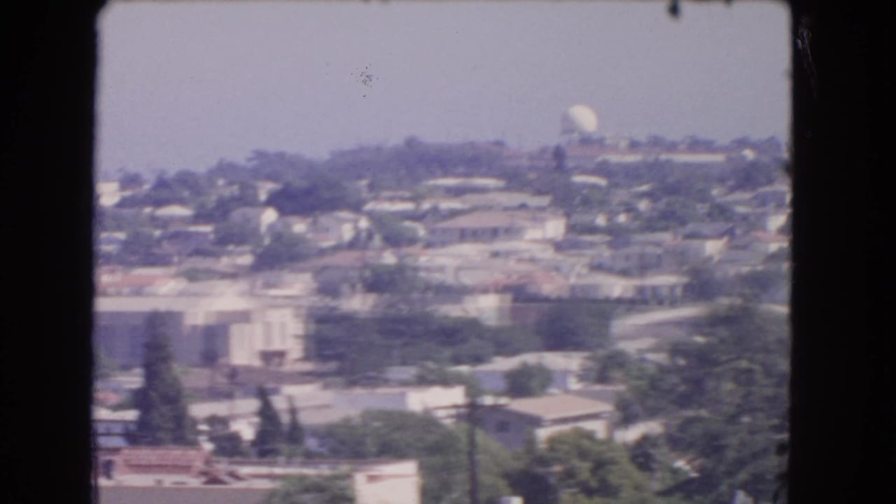 Hazy aerial view of hillside houses, trees, rooftops, and a distant white dome framed by dark film borders.